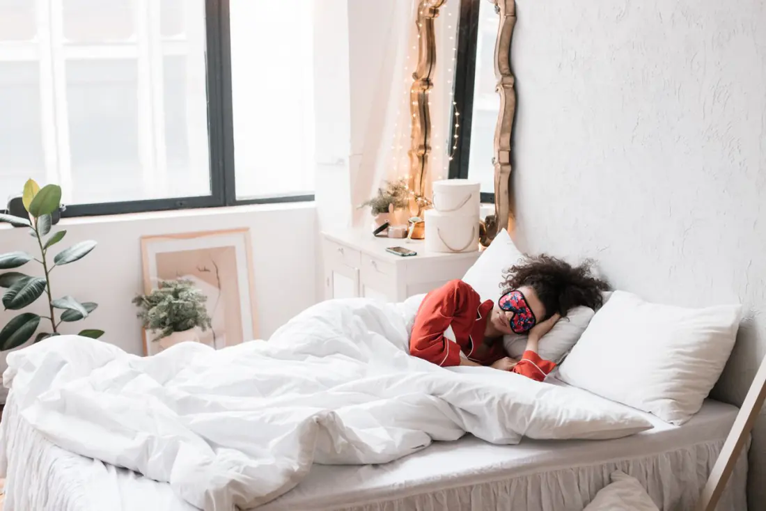 Calm, bright bedroom with plants and a woman resting with a sleep mask