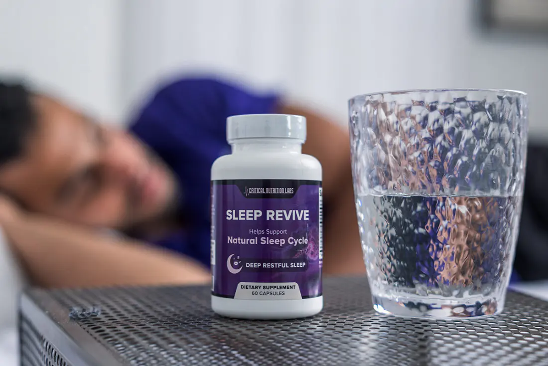 Sleep Revive bottle on a nightstand next to a glass of water, with a person sleeping in the background